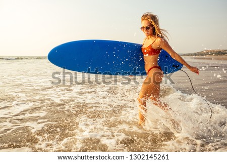beautiful young woman in orange bikini swimsuit with blue surfboard and shinning sun light run by paradise beach with water splash.happy girl waves, sand and surfing.concept of freedom and freelancing
