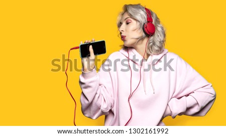 Portrait of charming happy woman holding modern device and wearing red headphones. Cute model singing in mobile phone and having fun. Isolated on yellow background
