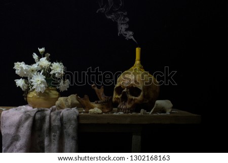 Skull wich has candle on head and white flower in vase and dirty cloth on wooden plank dim light , Still Life style / selective focus and space for texts

