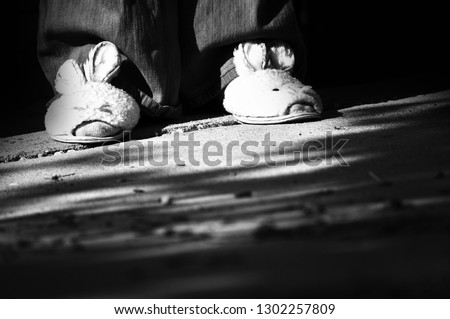 Black and white photo of girl's feet wearing sad bunny slipper house shoes sitting alone outside on concrete slab in a low income housing project one Easter afternoon.