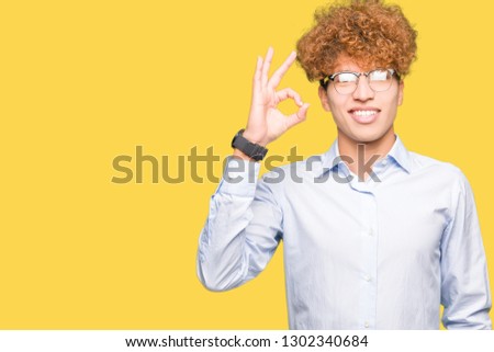 Young handsome business man with afro wearing glasses smiling positive doing ok sign with hand and fingers. Successful expression.