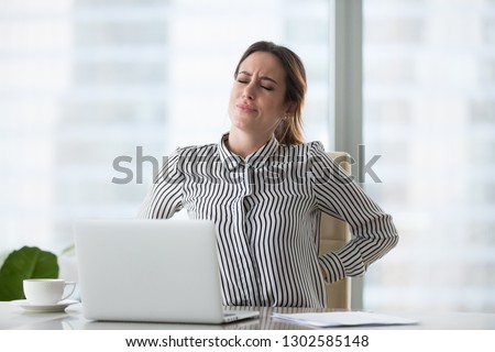 Tired fatigued businesswoman worker rubbing back feeling pain in kidneys suffers from lower lumbar backache after sedentary computer work sitting in incorrect posture on uncomfortable office chair Royalty-Free Stock Photo #1302585148