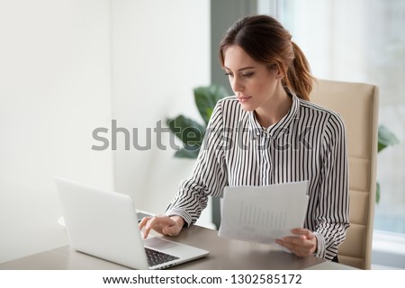 Serious focused businesswoman typing on laptop holding papers preparing report analyzing work results, female executive doing paperwork at workplace using computer online software for data analysis Royalty-Free Stock Photo #1302585172