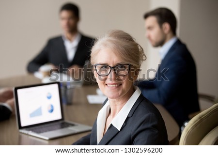 Smiling mature female company executive, professional manager, company ceo looking at camera, happy middle aged businesswoman in glasses posing at team office meeting, woman boss head shot portrait Royalty-Free Stock Photo #1302596200