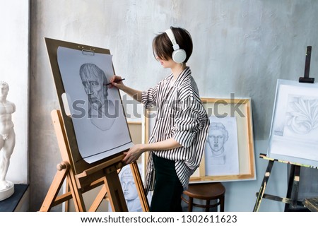Modern young freelancer artist sculptor woman in headphone creates new art masterpiece drawing sketch at art workshop workplace studio exhibition with drawings, sketches and gypsum plaster sculptures. Royalty-Free Stock Photo #1302611623