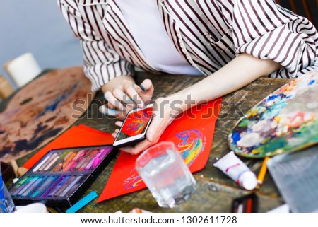 Soiled artists hands with smartphone make photo of pastel painting on orange paper. Creative workplace on wooden table with painting tools, pencils, pastels, tubes, paints, palette. Art concept.