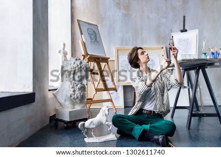 Young beautiful artist teacher student woman girl boy in modern art workshop studio surrounded by black white sketch painting big easel tools gypsum busts horse chooses stack of art brushes for oil 