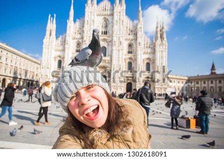 Winter travel, vacations and birds concept - Young happy woman tourist with funny pigeons making selfie photo in front of the famous Duomo cathedral in Milan.