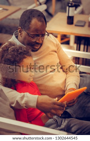 Watching cartoon. Loving caring father wearing glasses hugging his little princess watching cartoon