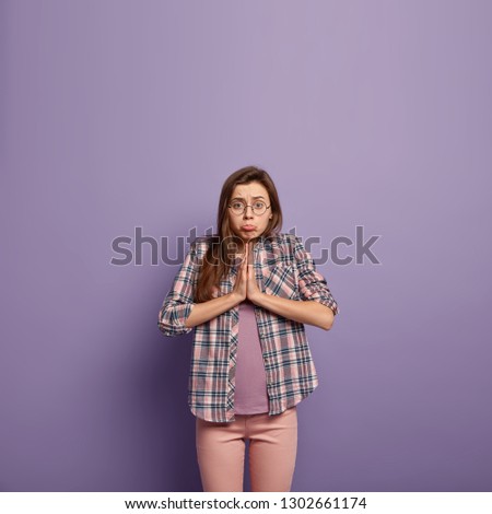 Miserable woman purses lower lips, pleads for mercy, has pity facial expression, keeps arms pressed together, wears spectacles and checkered shirt, isolated over purple wall with empty space