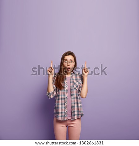 Surprised amazed beautiful woman with long dark hair, points with both fore fingers, exclaims with astonishment, dressed in checkered fashionable shirt and trousers, isolated on purple background