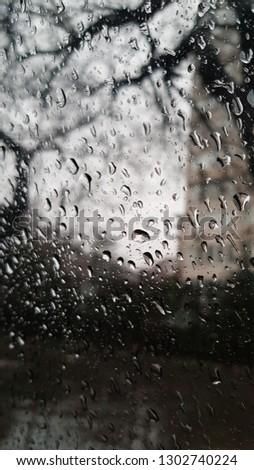 Raindrops are dripping on transparent glass window surface. Stormy weather. Rainy day background. Water drops texture. Dark wet backdrop with blurred tree silhouettes 