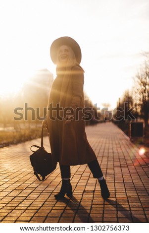 People, travel, holidays and adventure concept. Young woman with short loose hair walking on city street at sunset wearing hat and coat, enjoying happy pleasant moment of her vacations 