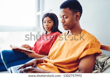 Serious african american woman looking at her boyfriend typing on laptop computer searching website for making online booking in bed room, dark skinned couple shopping in web store via netbook