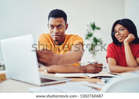 Serious african american hipster guy pointing on laptop computer browse information for doing homework with female colleague, dark skinned students watching tutorial while making coursework project