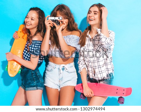 Three beautiful stylish smiling girls with colorful penny skateboards.Women in summer hipster checkered shirt clothes near blue wall. Taking pictures on retro camera