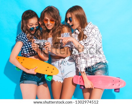 Three beautiful stylish smiling girls with penny skateboards.Women in summer hipster checkered shirt clothes and sunglasses near blue wall. Taking pictures on retro camera