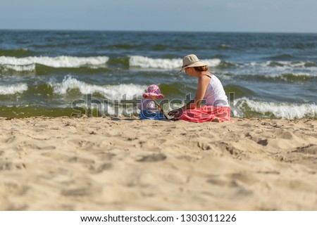 Mother with small baby girl relaxing on sea beach at sunny summer day.