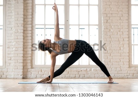 Young attractive yogi woman practicing yoga, doing Utthita Trikonasana exercise, extended triangle pose, working out, wearing sportswear, black pants and top, indoor full length, white yoga studio Royalty-Free Stock Photo #1303046143
