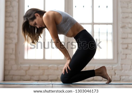 Young sporty attractive woman practicing yoga, doing Upward Abdominal Lock exercise, Uddiyana Bandha pose, working out, wearing sportswear, pants and top, indoor full length, white yoga studio