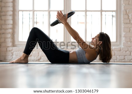 Young sporty attractive woman doing toning pilates exercise for abs with exercise circle, crunches for abdominal strengthening using pilates magic circle, wearing sportswear at yoga studio or at home Royalty-Free Stock Photo #1303046179