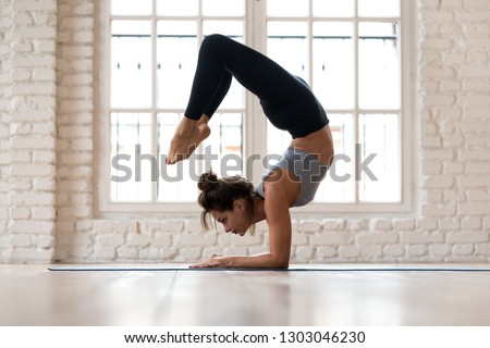 Young sporty attractive woman practicing yoga, doing Vrischikasana, Scorpion exercise, handstand pose, working out, wearing sportswear, black pants and top, indoor full length, yoga studio Royalty-Free Stock Photo #1303046230