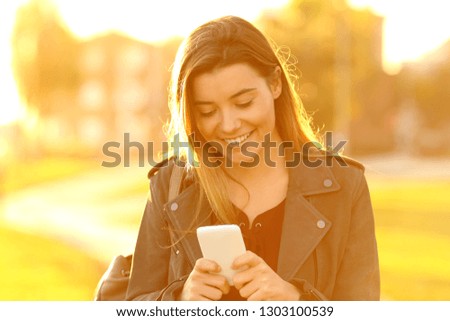 Front view portrait of a happy teenage girl at sunset using a smart phone in the street