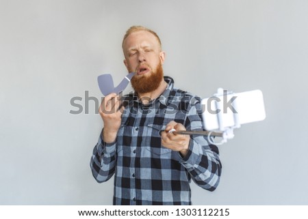 Red hair mature man standing isolated on grey wall holding monopod taking selfie photo on smartphone with paper tobacco pipe looking camera arrogantly