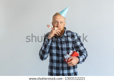 Red hair mature man wearing birthday cap standing isolated on grey wall holding gift box blowing into party horn looking camera smiling excited