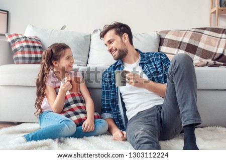 Bearded man and little girl at home family time sitting on carpet on floor holding cups drinking hot chocolate loooking at each other smiling joyful