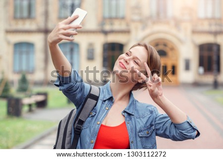 Young female student carrying backpack at university campus standing taking selfie photos on smartphone posing showing v sign looking camera smiling joyful