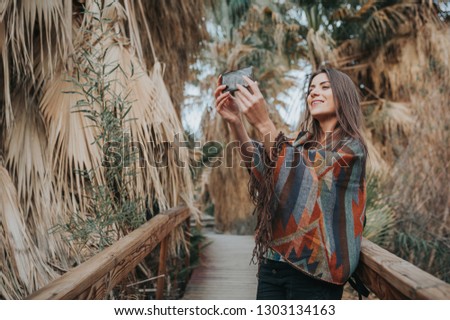 Smiling young woman holding smartphone, taking selfportraits outdoors, surrounded by palms.