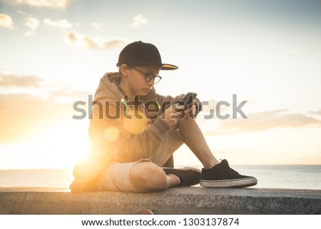 Handsome child with glasses and skater black hat sitting play with cellphone.Teen relaxing with phone enjoying outdoors at the sunset. Outdoor chatting with internet friends online near the ocean