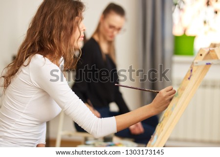 Girl with brown curly hair dressed in white blouse paints a picture at the easel in the drawing school