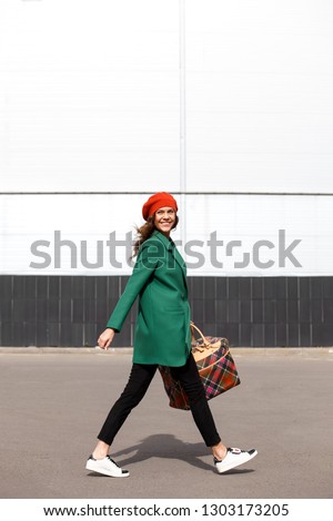 Charming stylish brunette girl dressed in a green coat and orange beret holding big checkered bag walks in the street on a sunny day