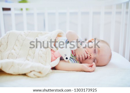 Adorable baby girl sleeping in co-sleeper crib attached to parents' bed with stuffed toy. Little child having a day nap in cot. Sleep training concept. Infant kid in sunny nursery Royalty-Free Stock Photo #1303173925