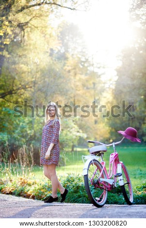 Happy blond fashionable long-haired attractive woman in short dress standing at lady bicycle on paved summer park alley on beautiful green and golden trees background in the evening.