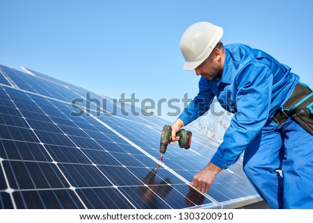Man worker in blue suit and protective helmet installing solar photovoltaic panel system using screwdriver. Professional electrician mounting blue solar module. Alternative energy ecological concept. Royalty-Free Stock Photo #1303200910