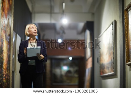 Portrait of mature woman holding clipboard looking at paintings standing in art gallery or museum, copy space Royalty-Free Stock Photo #1303281085