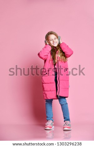full-length portrait of Cute smiling girl in winter puffer jacket and fur headphones over pink wall background