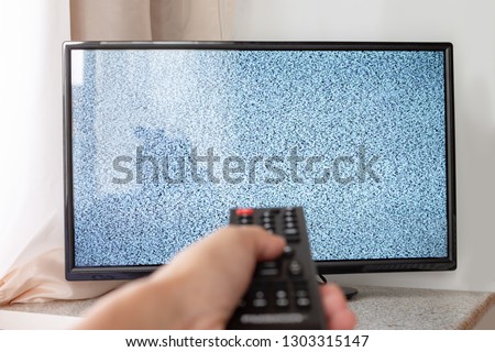 Hand with TV remote control in front of the screen with white noise on it - tuning the television channels and connecting problems.