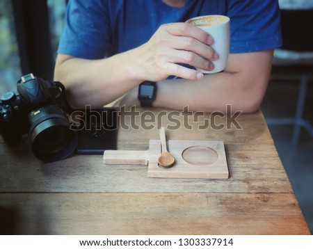 Photographer holding cup of hot coffee with DSLR camera and smartphone sitting in cafe shop. Hobby, vacation, Relax, chill time, traveler concept. Selective focus at hand