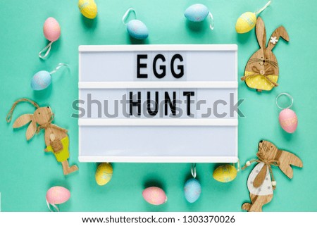 Egg hunt text on lightbox on green pastel paper background with yellow, pink, blue eggs  and bunnies. Bright template for Easter, top view, flat lay, holiday background with copy space 