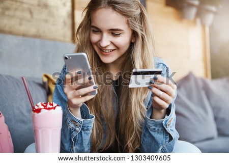 Carefree attractive fair-haired female friend in trendy denim jacket, holding credit card and smartphone, gazing at screen with smile while sending money to girlfriend who paid for drinks