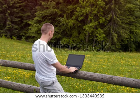 Man standing near the wooden fence in the field and works at a laptop near the pine forest.