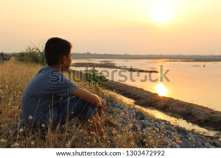 The boy looked at the sun in the evening by the river and was in a low light