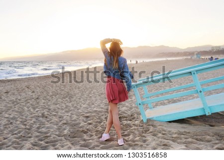 View from back of shapely beautiful traveling woman in   Pink dress, sneakers and jeans jacket looking on amazing sunset at California beach, ocean coast.