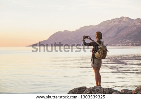 Young woman using smart phone taking photo of the sea at sunset, travel and active lifestyle concept