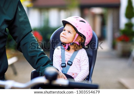 Portrait of little toddler girl with security helmet on the head sitting in bike seat and her father or mother with bicycle. Safe and child protection concept. Family and weekend activity trip. Royalty-Free Stock Photo #1303554679