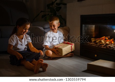 Little boy giving a giftbox with red ribbon to girl, siblings sitting near a fireplace